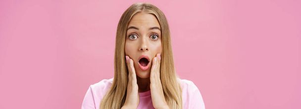 Close-up shot of shocked young female student with tanned skin and fair hair dropping jaw gasping from amazement touching cheeks surprised reacting to shocking stunning news over pink background. Copy - Photo, Image