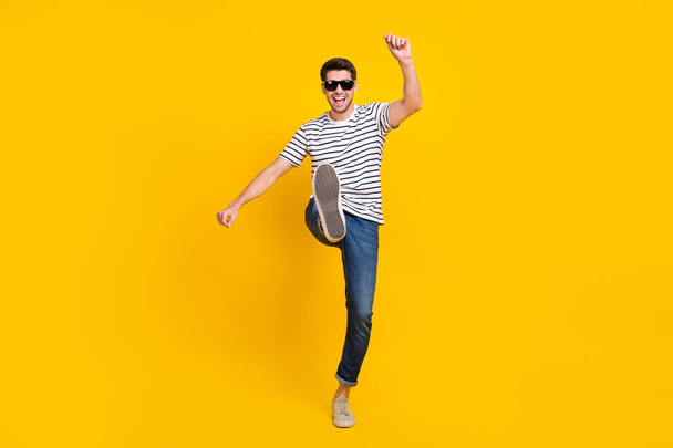 Full length photo of positive pretty guy dressed striped t-shirt dark eyewear walking having fun isolated yellow color background. - Zdjęcie, obraz