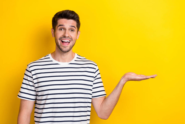 Photo of young excited surprised student man wear striped black white t-shirt hold palm copyspace object advert isolated on yellow color background. - Φωτογραφία, εικόνα