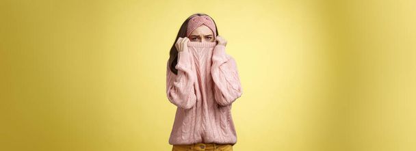 Sad silly, gloomy cute young girl hiding face in sweater collar pulling clothes on nose frowning displeased, unhappy, looking indignant and reluctant standing disappointed over yellow background. - Фото, зображення