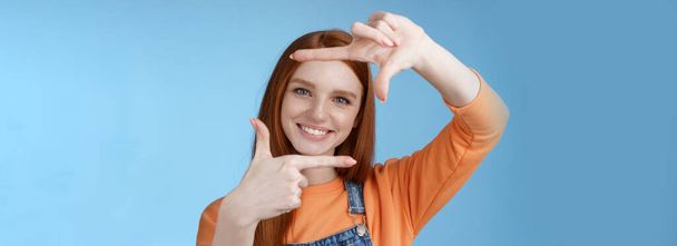 Kreative gut aussehende hübsche rothaarige weibliche Assistentin auf der Suche nach Inspiration lässt Handrahmen durch entzücktes Lächeln schauen, zufrieden gefundene großartige Aufnahme im Stehen, blauer Hintergrund. - Foto, Bild