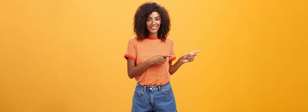 Çekici, arkadaş canlısı Afro-Amerikan kadın iş arkadaşının portresi solu işaret ediyor ve müşteriler için turuncu arka planda mükemmel bir kopya alanı gösteriyor. Yaşam biçimi. - Fotoğraf, Görsel