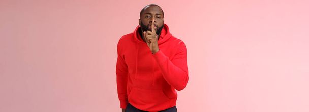 Shhh it taboo. Portrait sensual funny cute african-american bearded man in red hoodie bending camera show shush gesture index finger on lips telling secret asking keep quiet not tell anyone mystery. - Photo, Image