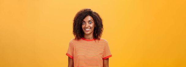 Waist-up πλάνο του κατάπληκτος και ενθουσιασμένος γοητευτικός Αφροαμερικανός γυναίκα με σγουρά μαλλιά σκάει τα μάτια από συγκίνηση και χαρά χαμογελώντας ευρέως είναι έκπληκτος από μεγάλο δώρο πάνω πορτοκαλί φόντο - Φωτογραφία, εικόνα