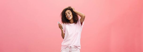 Ritratto di spensierata elegante moderna ragazza dalla pelle scura utilizzando smartphone appeso intorno gioiosamente toccando i capelli e guardando con ampio sorriso la fotocamera che tiene il cellulare in posa contro il muro rosa - Foto, immagini