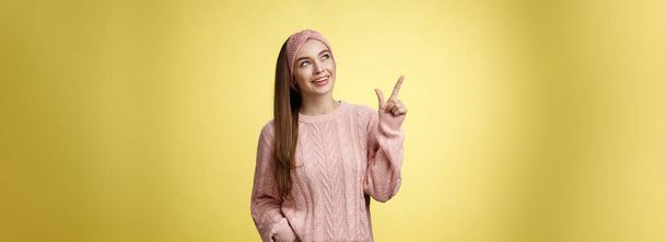 Portrait of amused interested cute 20s european girl wearing sweater, headband looking upper left corner pointing sideways smiling intrigued, ecstatic, captured by curious promo over yellow wall. - Photo, image