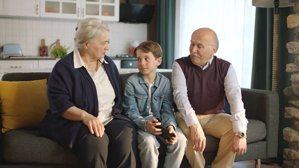 A kisfiú meglátogatta a nagyszüleit. Boldog idős pár ül a kanapén és beszélgetnek a kis unokájukkal.. - Fotó, kép