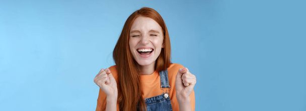 Lifestyle. Sincere happy rejocing ginger girl close eyes smiling broadly say yes waving clenched fists joyfully celebrate enterting university dream come true winning prize triumphing cheerfully blue - Photo, Image
