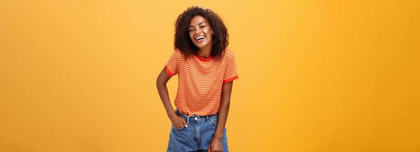 カリスマ的な魅力的なアフリカ系アメリカ人のスタイリッシュなアフリカ系アメリカ人女性のトレンディーなショートパンツとTシャツの肖像が喜んでオレンジの背景の上にポーズを笑ってクールな人々と話を楽しんで笑って - 写真・画像