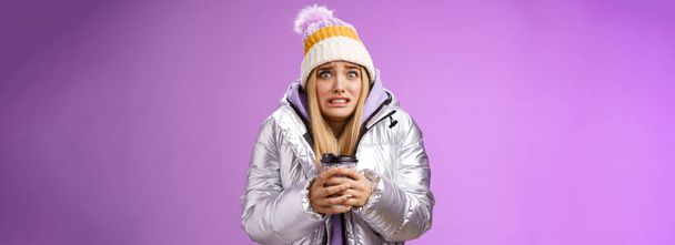 Shaking uncomfortable displeased girl wearing stylish silver jacket hat trembling freezing cold warm hands holding take-away coffee cup clench teeth pop eyes discomfort, standing purple background. - Photo, image
