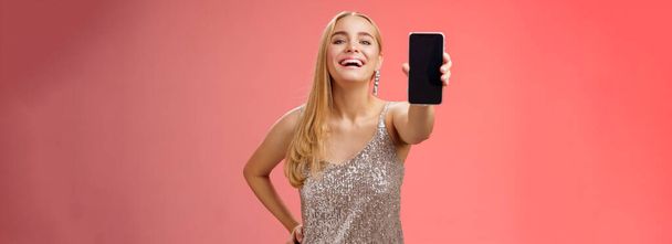 Dumny radosny uroczy blond europei kobieta w stylowej srebrnej lśniącej sukience trzymać talię pewnie przedłużyć ramię pokazując wyświetlacz smartfona niesamowite nowe urządzenie aplikacji, czerwone tło. - Zdjęcie, obraz