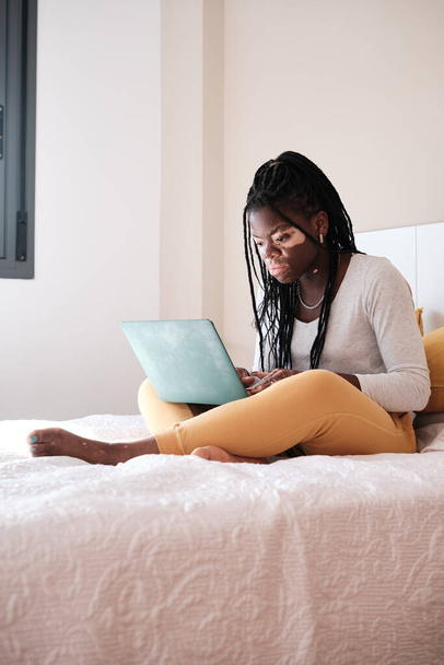 Volledig lichaam van jonge vrouwelijke freelancer in casual kleding zitten op bed en browsen laptop tijdens het werken aan remote project thuis - Foto, afbeelding