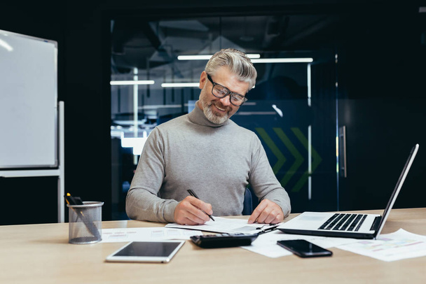 Senior successful businessman and investor working on paper work in the office, mature gray haired male boss working with contracts and accounts financier using laptop at work. - Foto, afbeelding