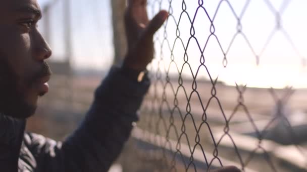 Male prisoner looking through a wire fence thinking about life and showing regret about bad choices. Young african man worried about future behind bars outside in a prison yard after doing crime. - Materiał filmowy, wideo