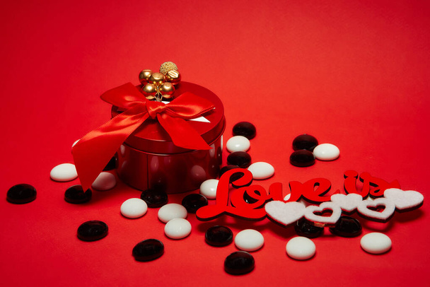Czerwone okrągłe pudełko z łukiem i dekoracji na górze, napis miłość jest z białymi sercami, białe i czarne okrągłe błyszczące kamyki na czerwonym tle. - Zdjęcie, obraz