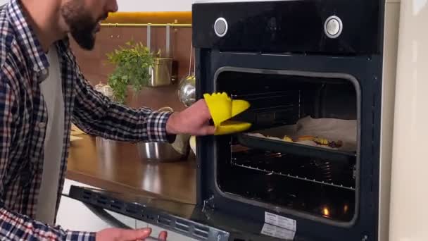 Closeup pozitivní muž vaření pomocí trouby v kuchyni pro domácnost. Veselý domácí manžel v rukavici, otvírající sporák, ochutnávající jídlo na pánvi, uspokojující přikyvování. Muž připravuje večeři uvnitř - Záběry, video
