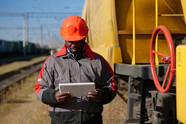 Inspektor vozů na nákladním nádraží se podívá do svého tabletu. Africká americká železnice muž s tabletem PC na nákladní vlak terminálu - Fotografie, Obrázek