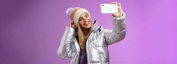 Веселая очаровательная девушка, весело проводящая время, выкладывает фотографии с отдыха на горнолыжном курорте, держа в руках смартфон, мигает языком, выглядит мобильным дисплеем, на фиолетовом фоне. - Фото, изображение