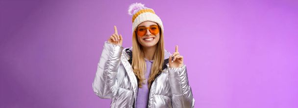 Lebensstil. Fröhlich energetisiert unterhalten niedlichen blonden Frau Spaß genießen Urlaub schneebedeckten Bergtour mit Sonnenbrille silberne Jacke Wintermütze tanzen zeigt amüsiert stehend lila Hintergrund - Foto, Bild