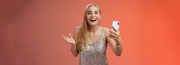 Astonished happy charming blond girl in silver glittering stylish dress holding smartphone amazed liking awesome result edit photo app smiling wondered amused, standing red background. - 写真・画像