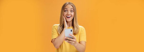 Ritratto di donna sorpresa e impressionata che reagisce all'app impressionante nello smartphone toccando la guancia dallo stupore e dalla gioia sorridendo ampiamente alla fotocamera che tiene il cellulare in mano sulla parete arancione. Tecnologia - Foto, immagini