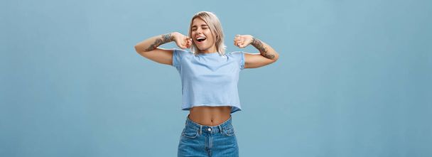 Great feelings after nap. Satisfied carefree and relaxed attractive athletic and stylish female with tattooed arms stretching hands yawning and smiling with closed eyes and pleased look after sleep - Photo, Image