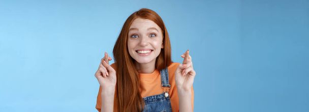 Izgatott érzelmi boldog vidám vörös hajú lány mosolygó optimista bámulás meglepett izgatott kereszt ujjak jó szerencse hiszem álom valóra kívánság előre csak a jó hír, kék háttér. - Fotó, kép