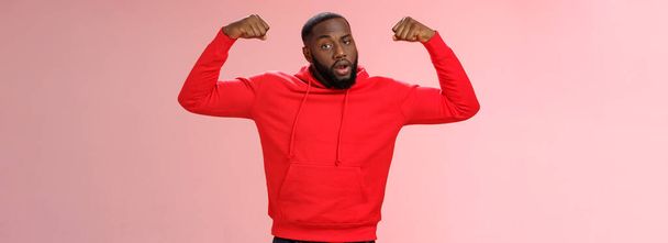 Сильні щокуваті афро-американські спортсмени з серйозним виглядом в червоному вихвалянні копит піднімають руки, показуючи великі м'язи біцепсів, стоячи впевнено рожевого фону. Копіювальний простір - Фото, зображення