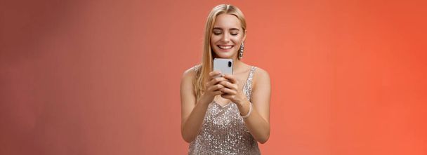 Entzückte zarte Glamour-Blondine in silbernem stylischem Glitzerkleid brillant und Ohrringe in der Hand, die ein Smartphone hält, mit dem ein Freund Moment der Feier festhält, roter Hintergrund lächelnd. - Foto, Bild