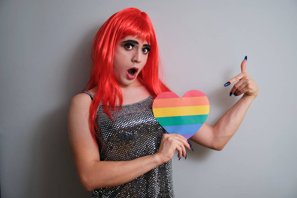 Ritratto di uomo fluido di genere sorpreso e con un cuore di bandiera arcobaleno in mano. queer LGBTQ. - Foto, immagini