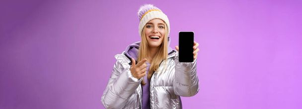 Tevreden geamuseerd goed uitziende blonde meisje suggereren neem kijkje smartphone display glimlachen gelukkig wijzen mobiele telefoon blij praten over geweldige nieuwe app functies, staande paarse achtergrond. - Foto, afbeelding