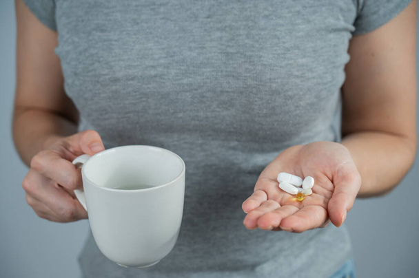 Faceless woman holding handful of pills and mug on gray background - Fotó, kép