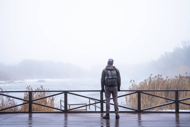 Молодой человек в тумане стоит один на деревянном пешеходном мосту и смотрит на озеро. Парень в капюшоне. Мирная атмосфера в природе. Наслаждаюсь свежим воздухом зимой. Вид сзади. - Фото, изображение