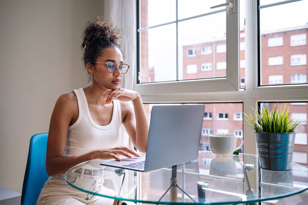 Молода афроамериканська жінка в повсякденному одязі з кучерявим волоссям торкається обличчя і переглядає ноутбук, сидячи за столом біля вікна і працюючи вдома над вільним проектом. - Фото, зображення