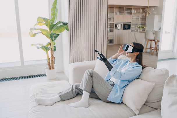 Disabled girl with artificial limb wearing virtual reality glasses learning to use bionic prosthetic arm during rehabilitation after limb loss, sitting on sofa in cozy living room. Medical high tech. - Photo, Image