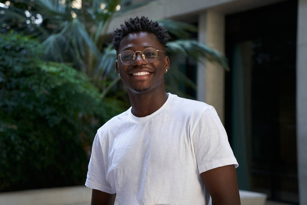 Portrait of black man very happy, smiling and looking at camera. An African American student sincerely smiles while standing outdoors. The guy is wearing a white t-shirt - Foto, Bild