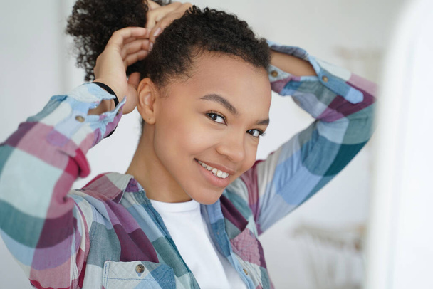 Glimlachend jong gemengd ras meisje met gezonde sterke afro haren, doen kapsel, gelukkig tevreden mooie tiener dame kijkt in spiegel maakt paardenstaart. Natuurlijke schoonheid, zelfzorg routine concept. - Foto, afbeelding