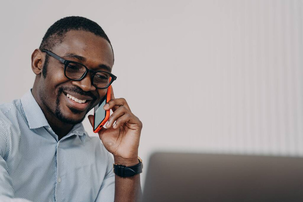 Φιλικός χαμογελαστός Αφροαμερικάνος επιχειρηματίας απαντά σε τηλεφωνική επαγγελματική κλήση κοιτάζοντας την οθόνη του φορητού υπολογιστή. Ευτυχισμένο μαύρο αρσενικό μάρκετινγκ και πωλήσεων διευθυντής συνομιλίες, συμβουλευτικός πελάτης, απολαμβάνοντας κινητή συνομιλία. - Φωτογραφία, εικόνα