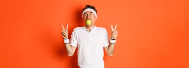 Portret zabawnego sportowca w średnim wieku, trzymającego jabłko w ustach i pokazującego znaki pokoju, stojącego nad pomarańczowym tłem w mundurze treningowym. - Zdjęcie, obraz