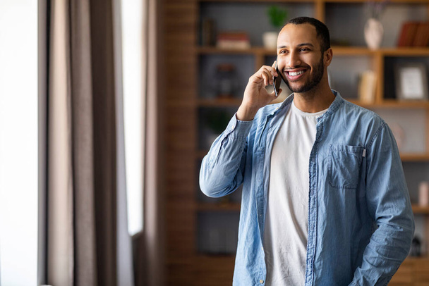 Portrait d'un bel homme noir souriant parlant sur un téléphone portable à la maison, jeune homme afro-américain joyeux debout près de la fenêtre dans le salon et bénéficiant d'une conversation mobile agréable, espace de copie - Photo, image