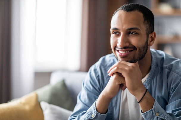 Smiling Handsome Black Guy Sitting On Sofa At Home And Looking Away, Pensive Young African American Man Resting Chin On Hands While Daydreaming In Living Room Interior, Closeup Shot With Copy Space - Photo, Image