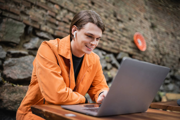 Lachende freelancer in oortelefoon met behulp van laptop in outdoor cafe  - Foto, afbeelding
