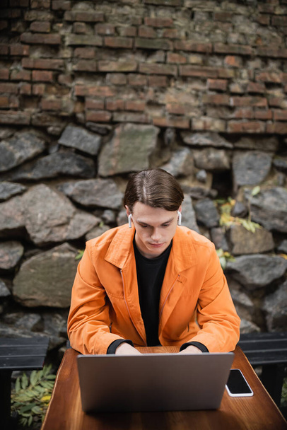 Fiatal férfi fülhallgató segítségével laptop közelében okostelefon üres képernyő szabadtéri kávézóban  - Fotó, kép