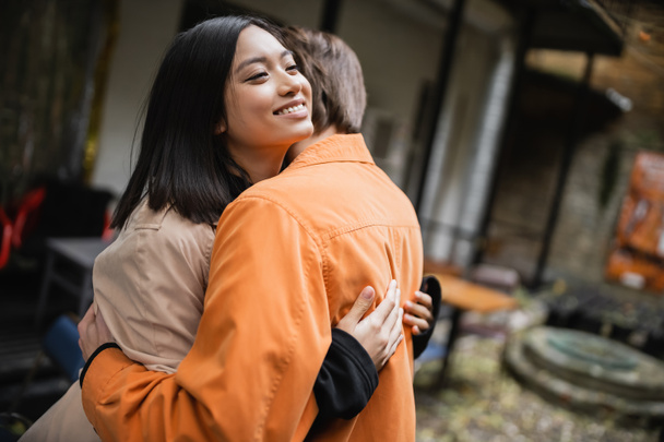 Sonriente mujer asiática en gabardina abrazando novio en terraza de café  - Foto, imagen