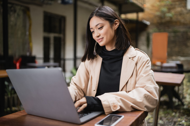 Positive asiatische Freelancer mit Laptop in der Nähe von Smartphone in Outdoor-Café  - Foto, Bild