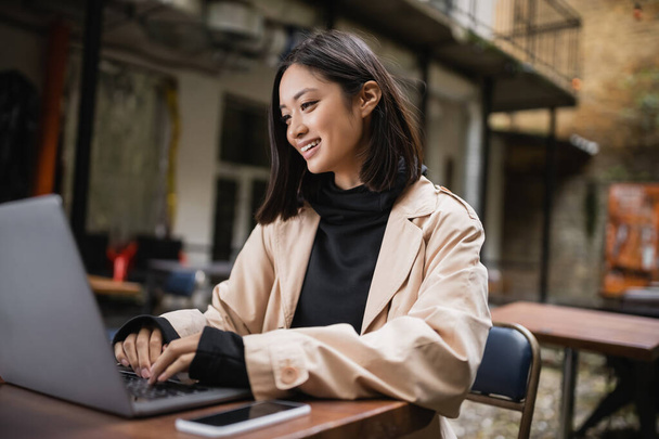 Alegre blogger asiático en gabardina usando laptop en terraza de café  - Foto, imagen