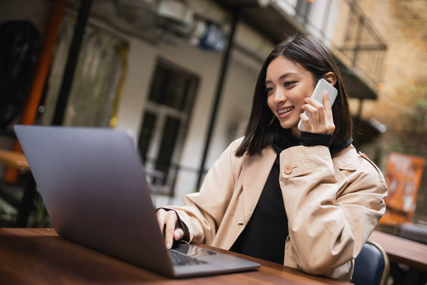 Positive asian freelancer talking on smartphone near blurred laptop in outdoor cafe  - 写真・画像
