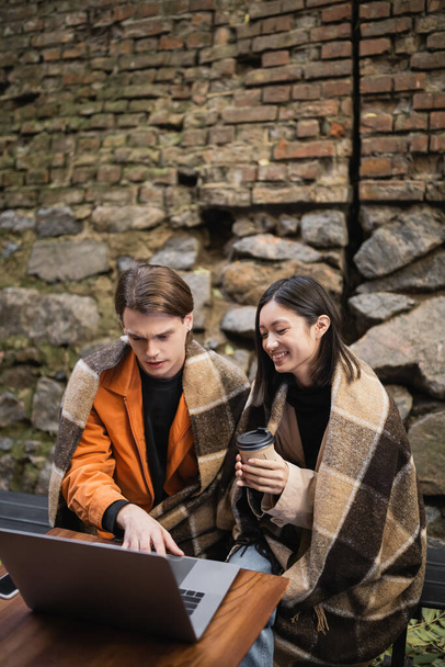 Cheerful asian woman holding coffee to go near boyfriend using laptop on terrace of cafe  - Photo, image
