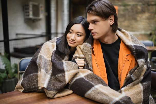 Smiling asian woman in blanket hugging boyfriend on terrace of cafe  - Photo, Image
