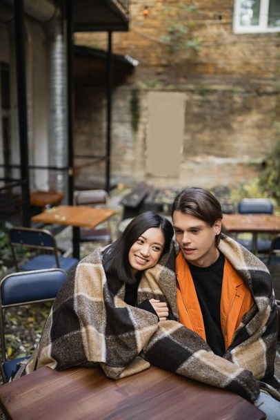 Cheerful asian woman in blanket hugging boyfriend on terrace of cafe  - Zdjęcie, obraz